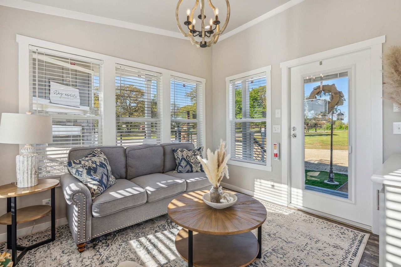 Bright sunroom with gray sofa, round coffee table, bay windows, and glass door overlooking a garden.