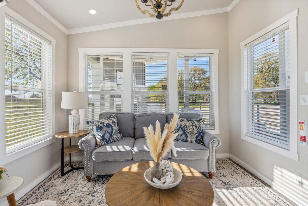 Bright sunroom with gray sofa, round wood table, large windows, and striped blinds