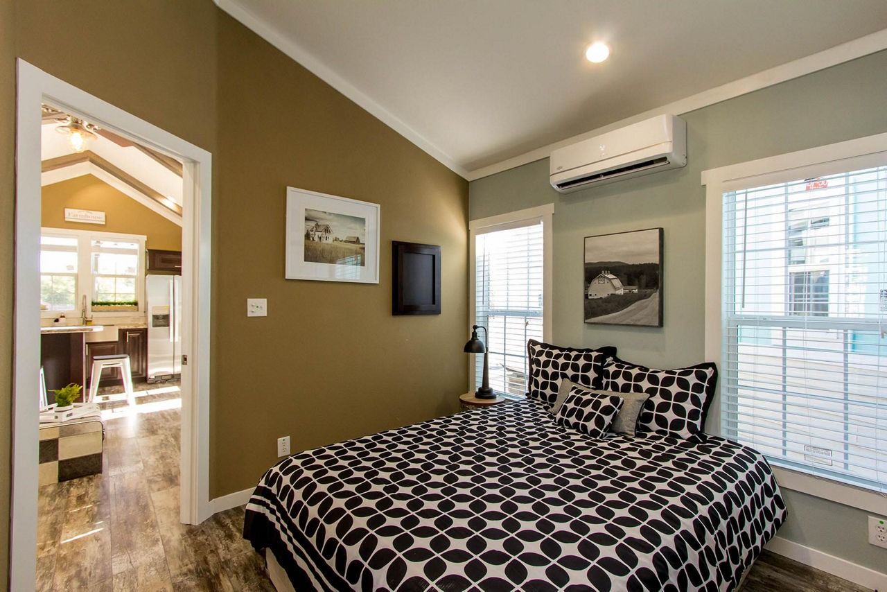Bedroom with patterned bedspread, wall-mounted TV, windows, and doorway to a bright adjacent room
