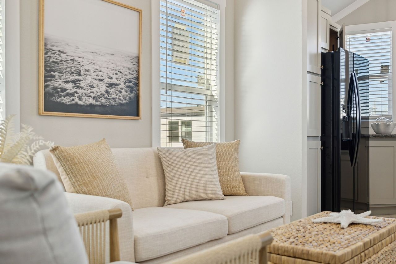 Bright living room with cream sofa, woven ottoman, and framed coastal art by tall windows