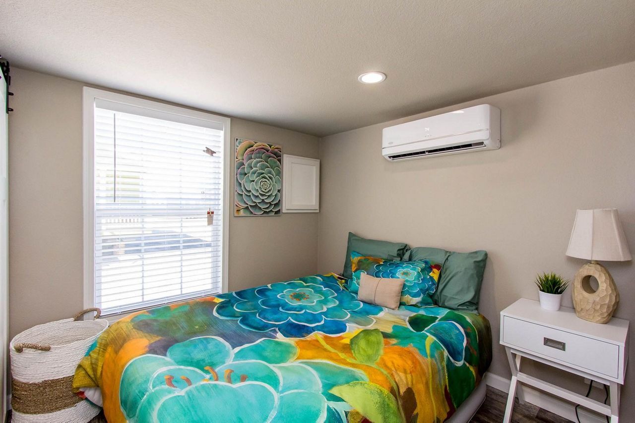 Cozy bedroom with turquoise and yellow bedspread, window blinds, nightstand, lamp, and wall-mounted AC unit