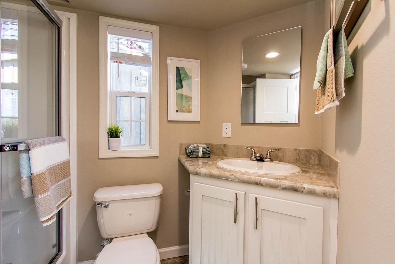 Small beige bathroom with toilet, vanity sink, mirror, window, and towel rack.