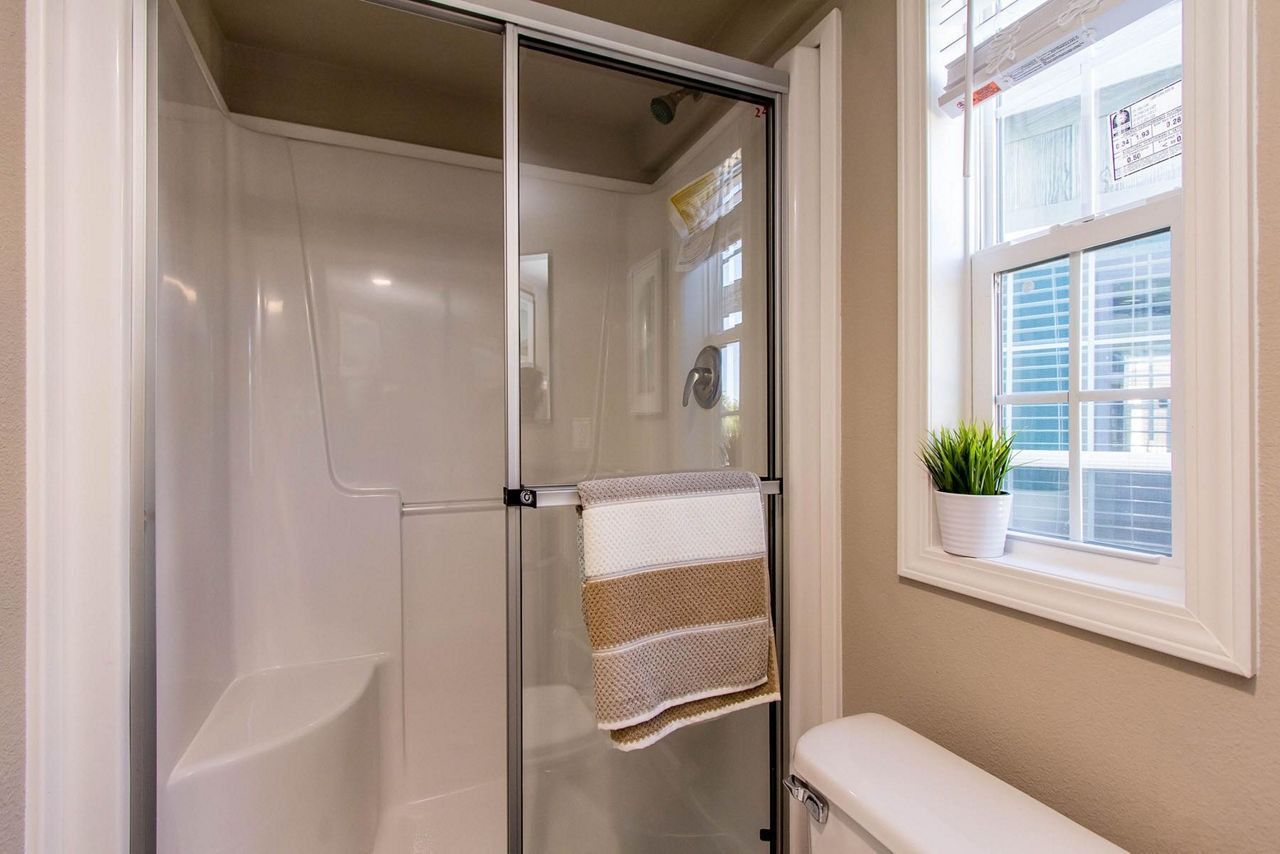 Bathroom shower with glass door, white tile, towel rack, and window beside a toilet