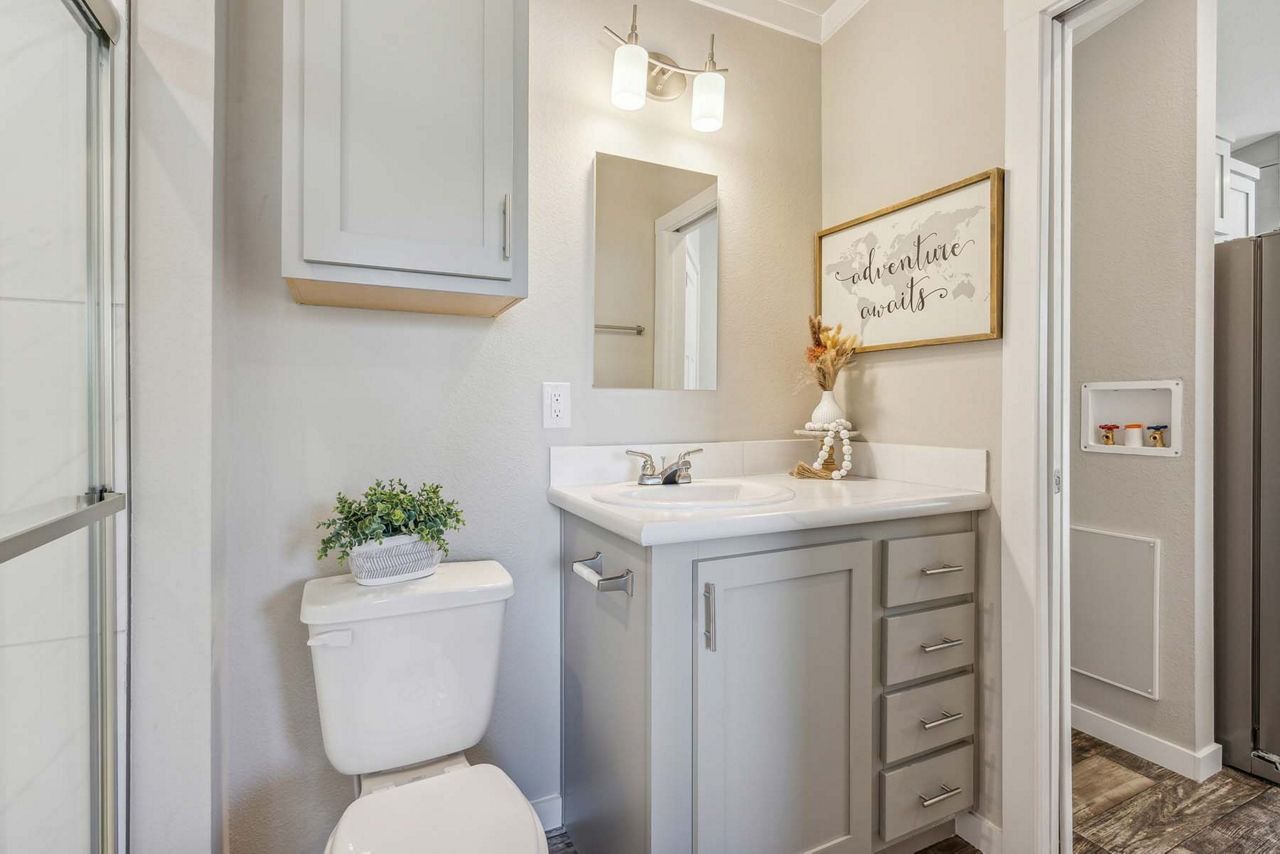Small bathroom with gray vanity, white toilet, mirror, and beige walls with decor.