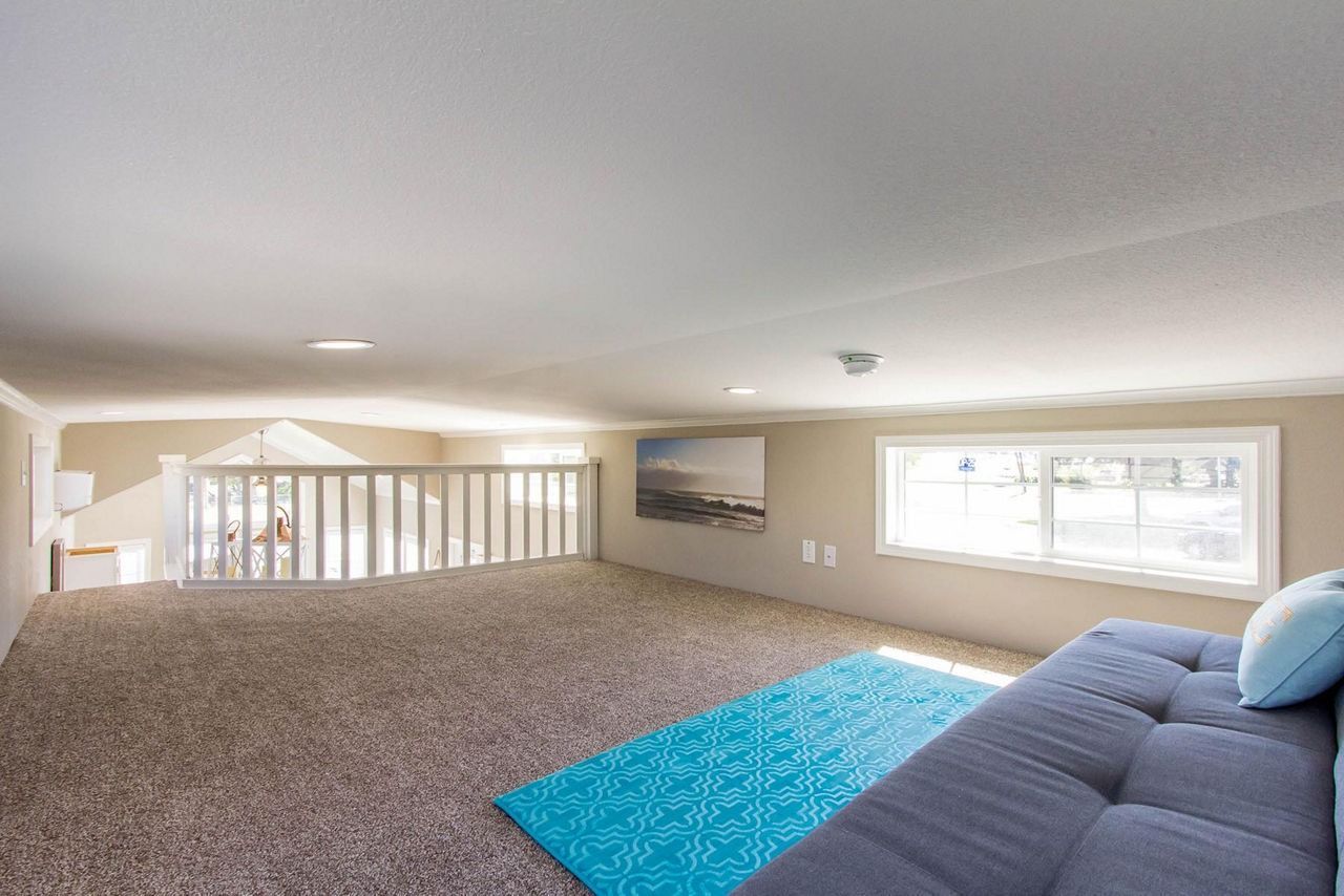 Empty carpeted living room with gray sofa, blue rug, and large window overlooking railing.