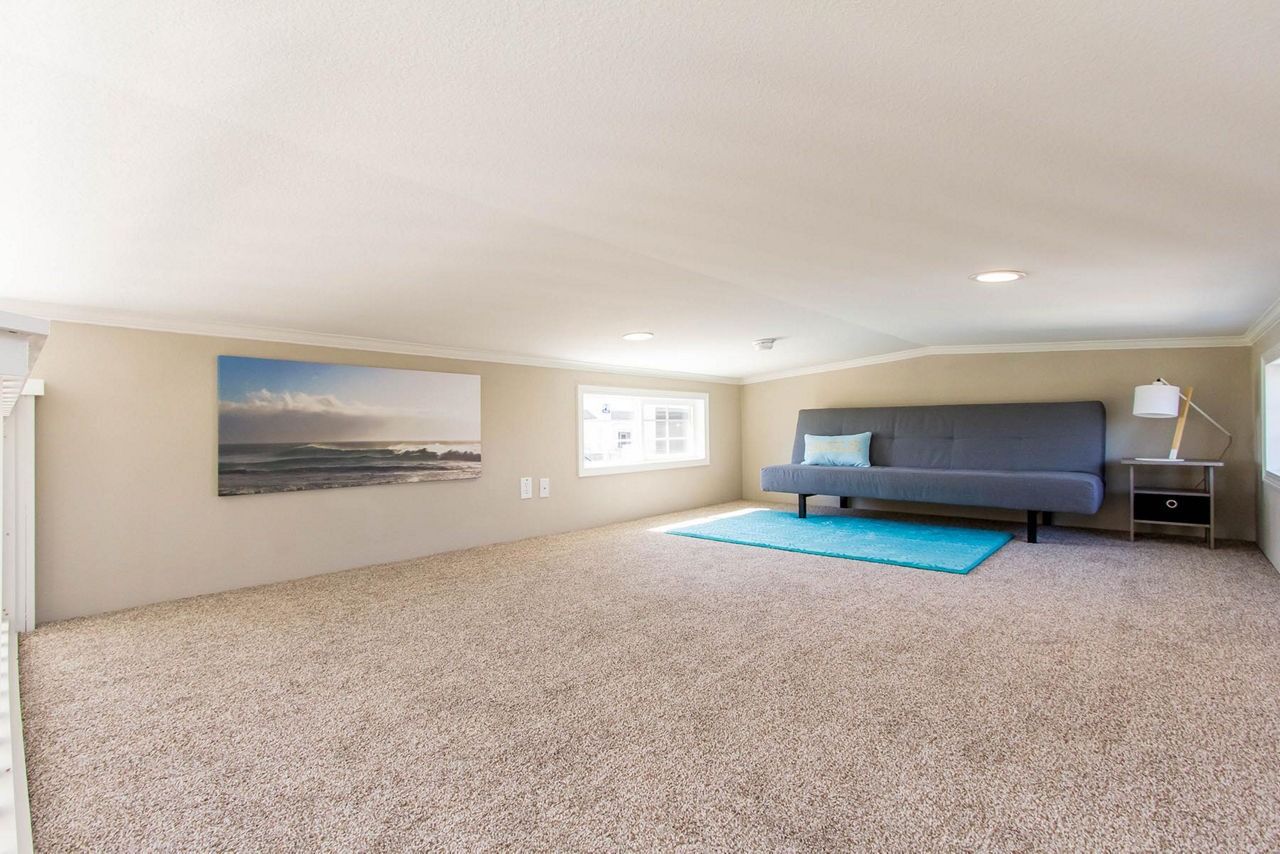Spacious carpeted bedroom with a gray bed, blue rug, wall art, and a small window.
