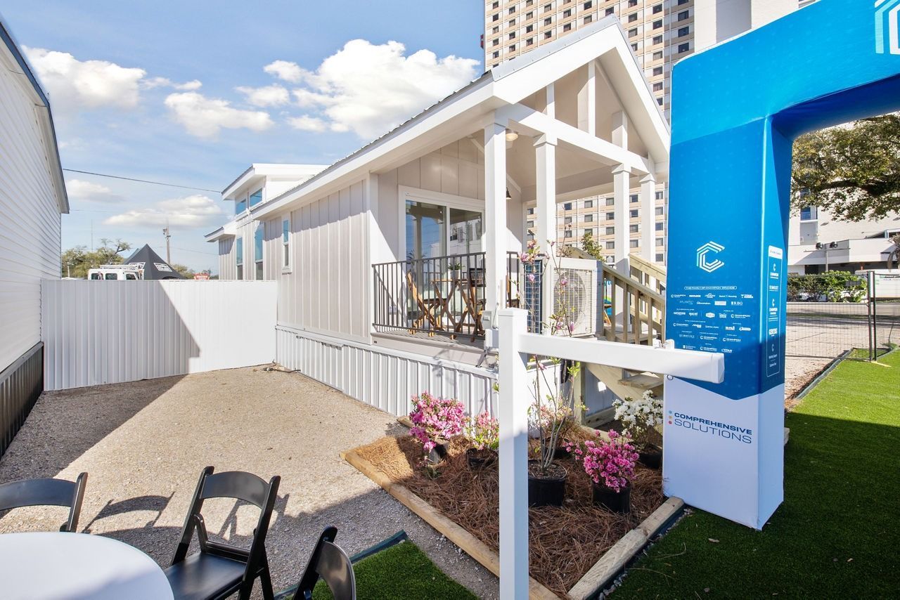 Outdoor white tiny house display with porch, blue sign, and city buildings in the background