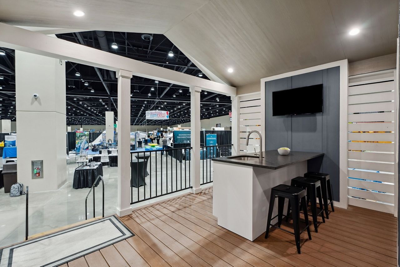 Modern trade show booth with white walls, bar counter, stools, and a wall-mounted TV in a bright exhibit hall