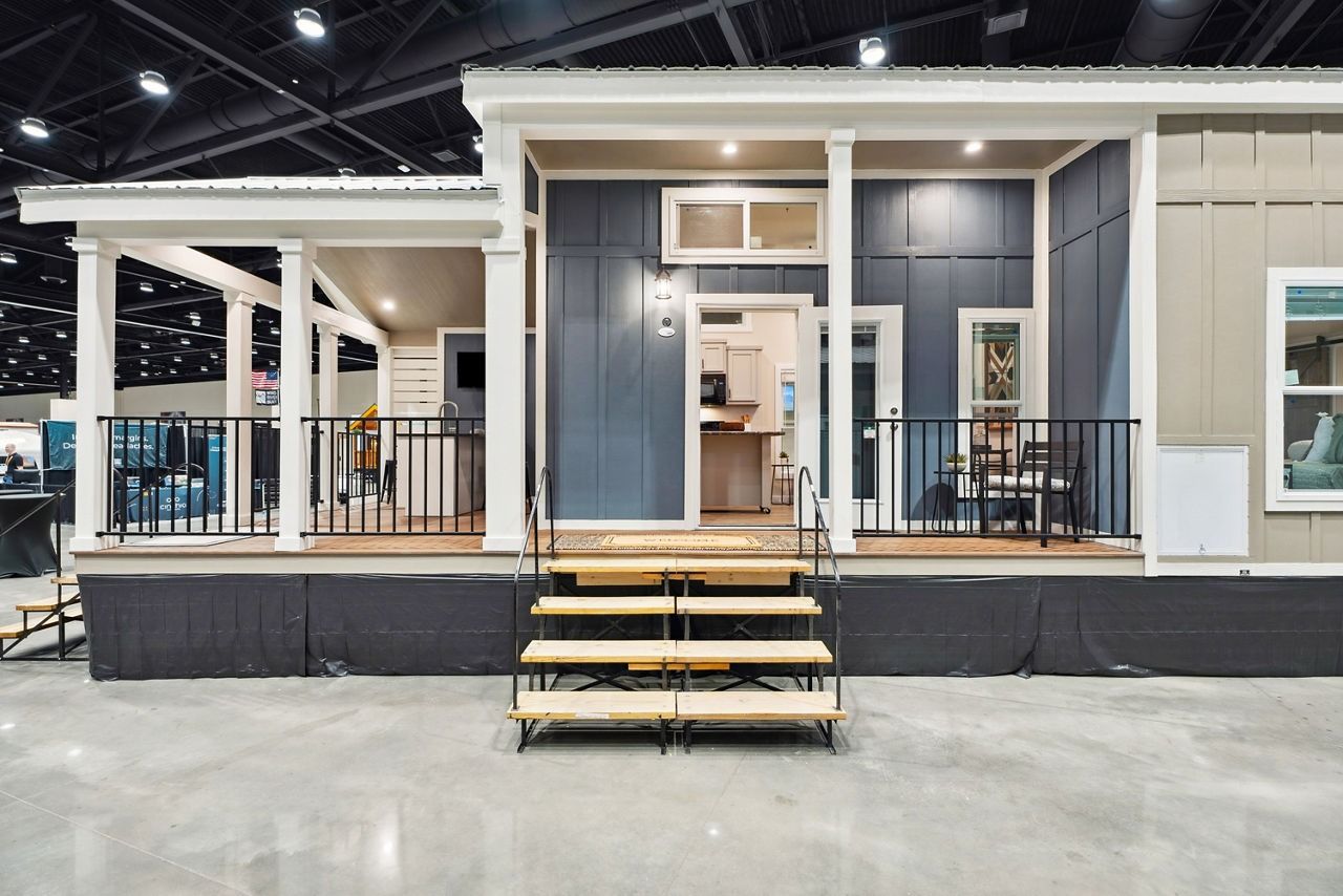 Modern tiny house display with front porch, steps, and black trim inside an exhibition hall