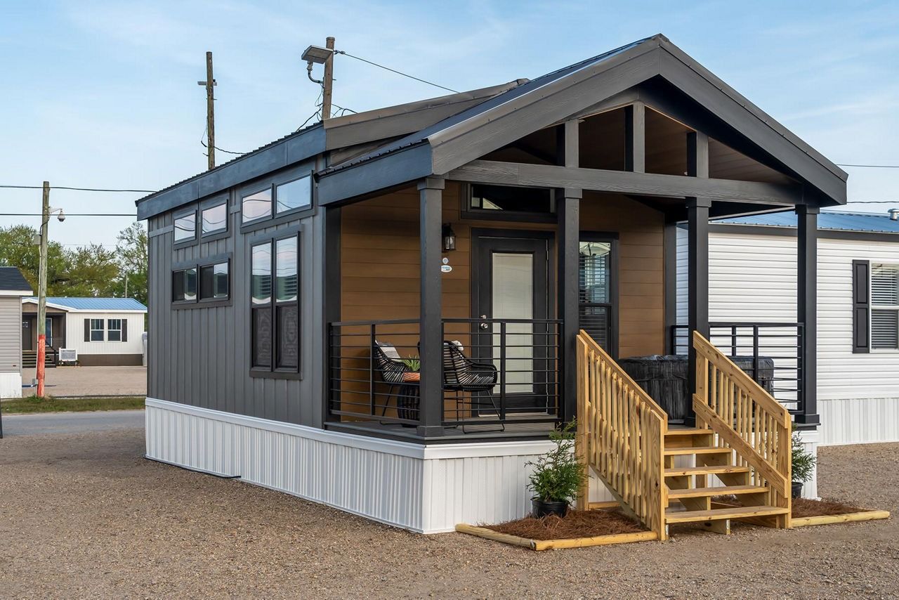 Small gray and white tiny house with porch steps and wooden railings on a gravel lot