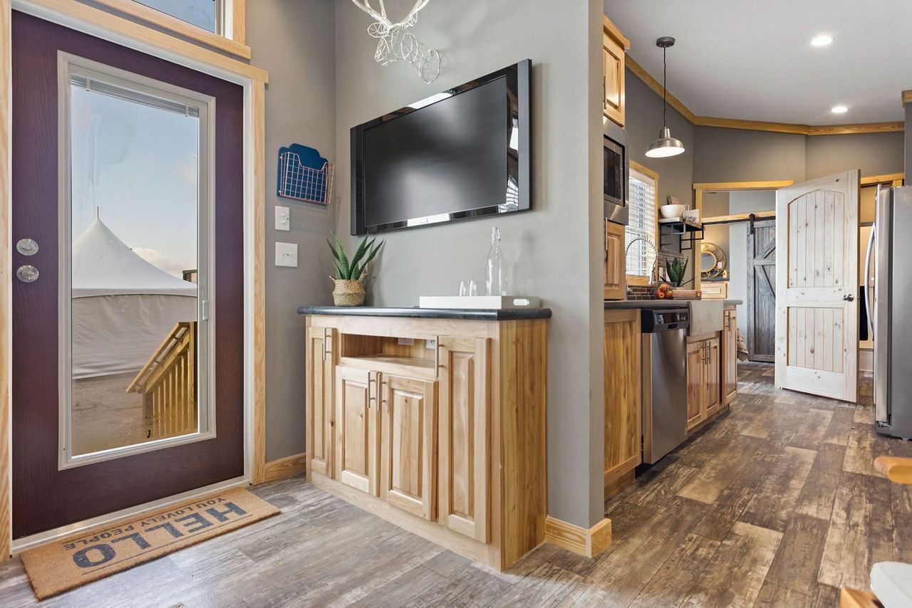 Modern hallway with a glass door, wall-mounted TV, wood cabinet, and open kitchen with wood floors.