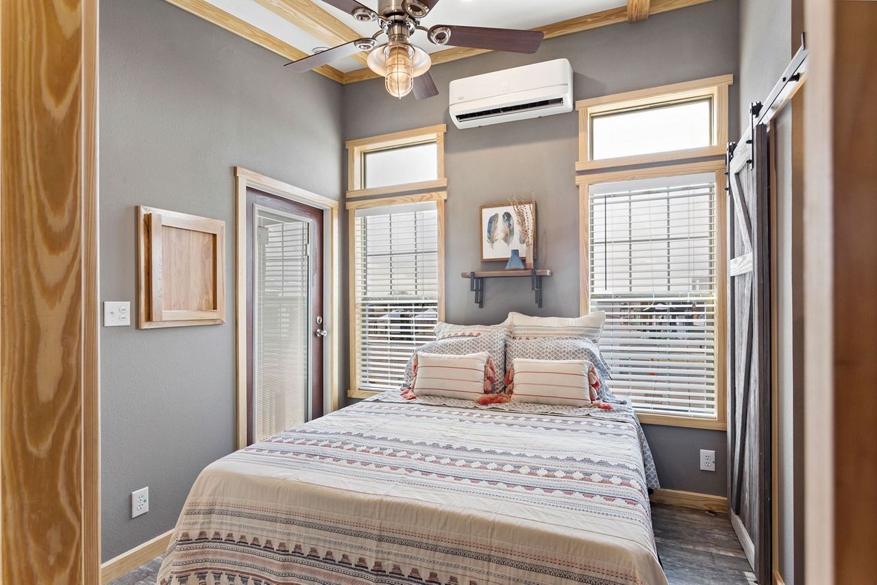 Bright bedroom with a bed, gray walls, wood trim, blinds, ceiling fan, and an air conditioner.