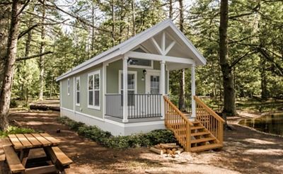 Small white cabin with a front porch and steps in a wooded forest setting.
