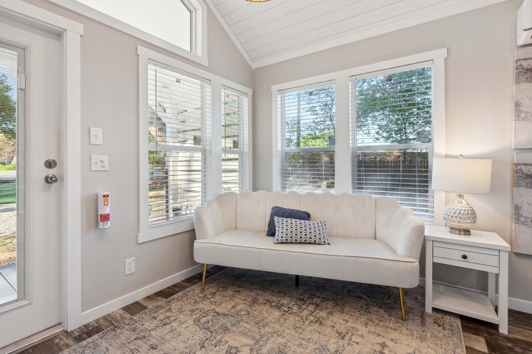 Bright living room with a white loveseat, large windows, patterned rug, and a side table with lamp.