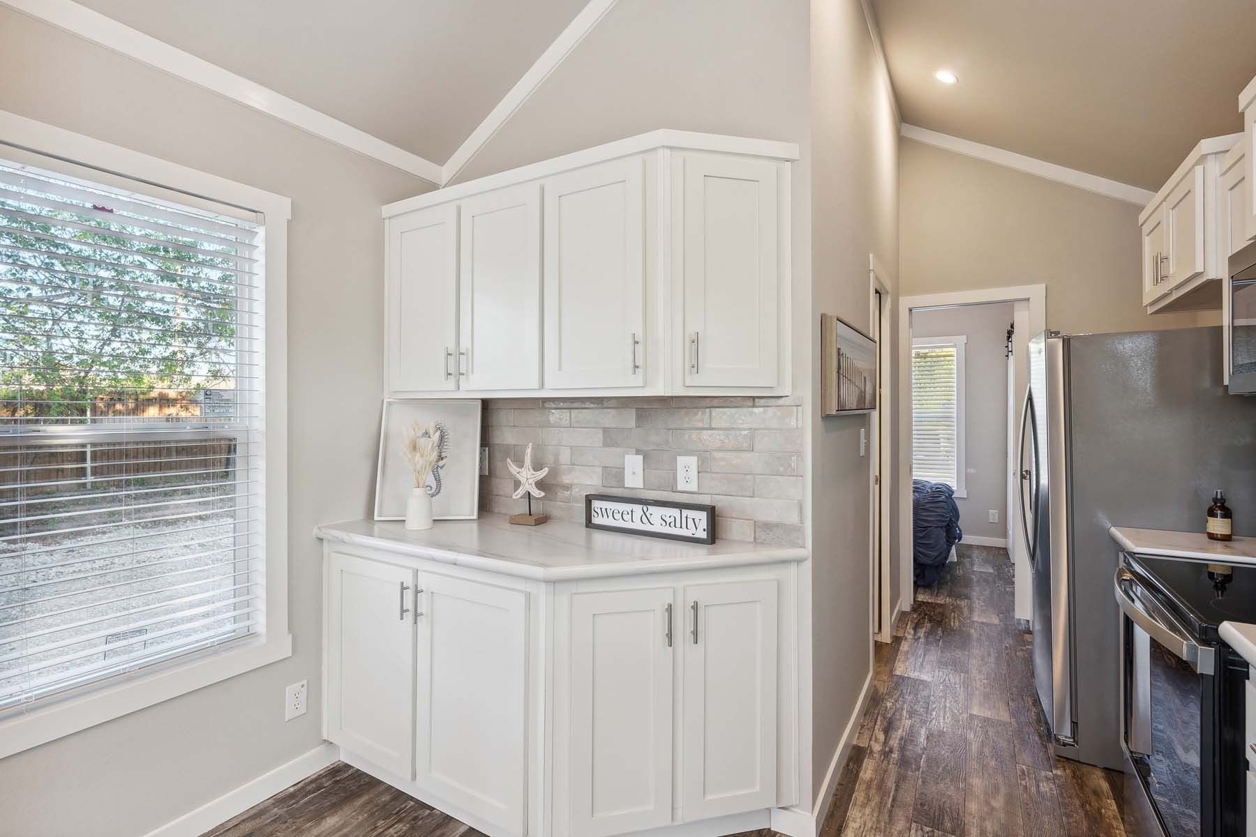 Bright kitchen with white cabinets, stainless stove, tile backsplash, and a hallway leading to another room.