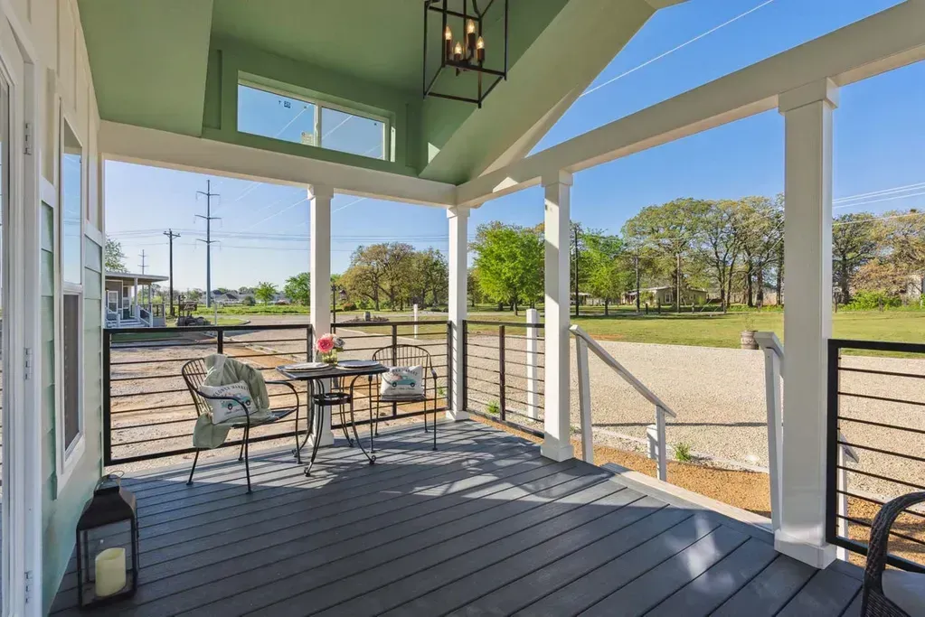 A covered outdoor deck with dark gray wooden flooring, a small bistro table with two chairs, and light green ceilings.