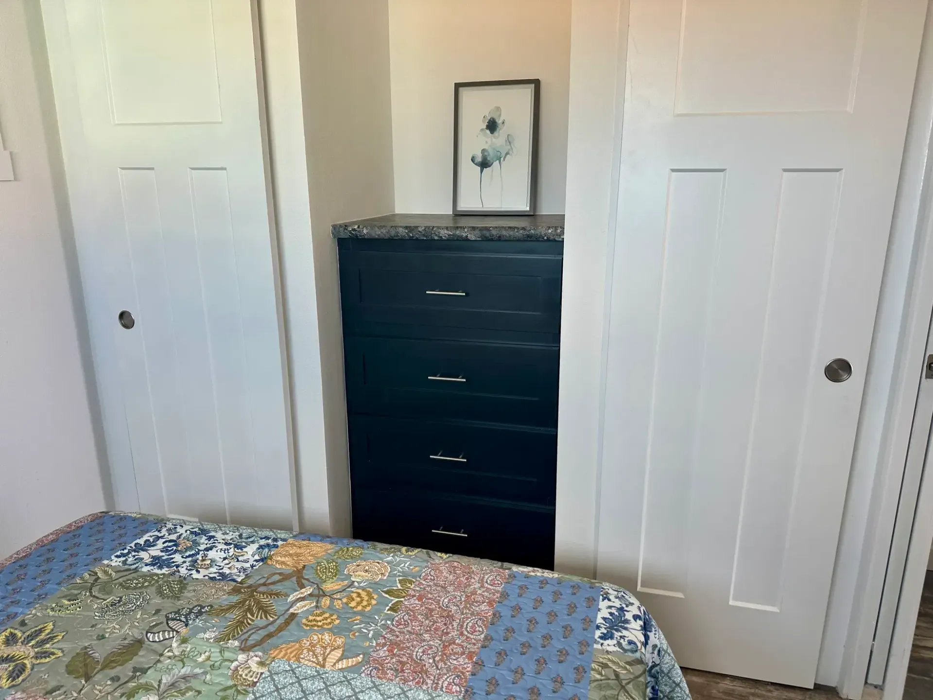 A black dresser sits between two white closet doors, topped with a small framed picture of a floral sketch.