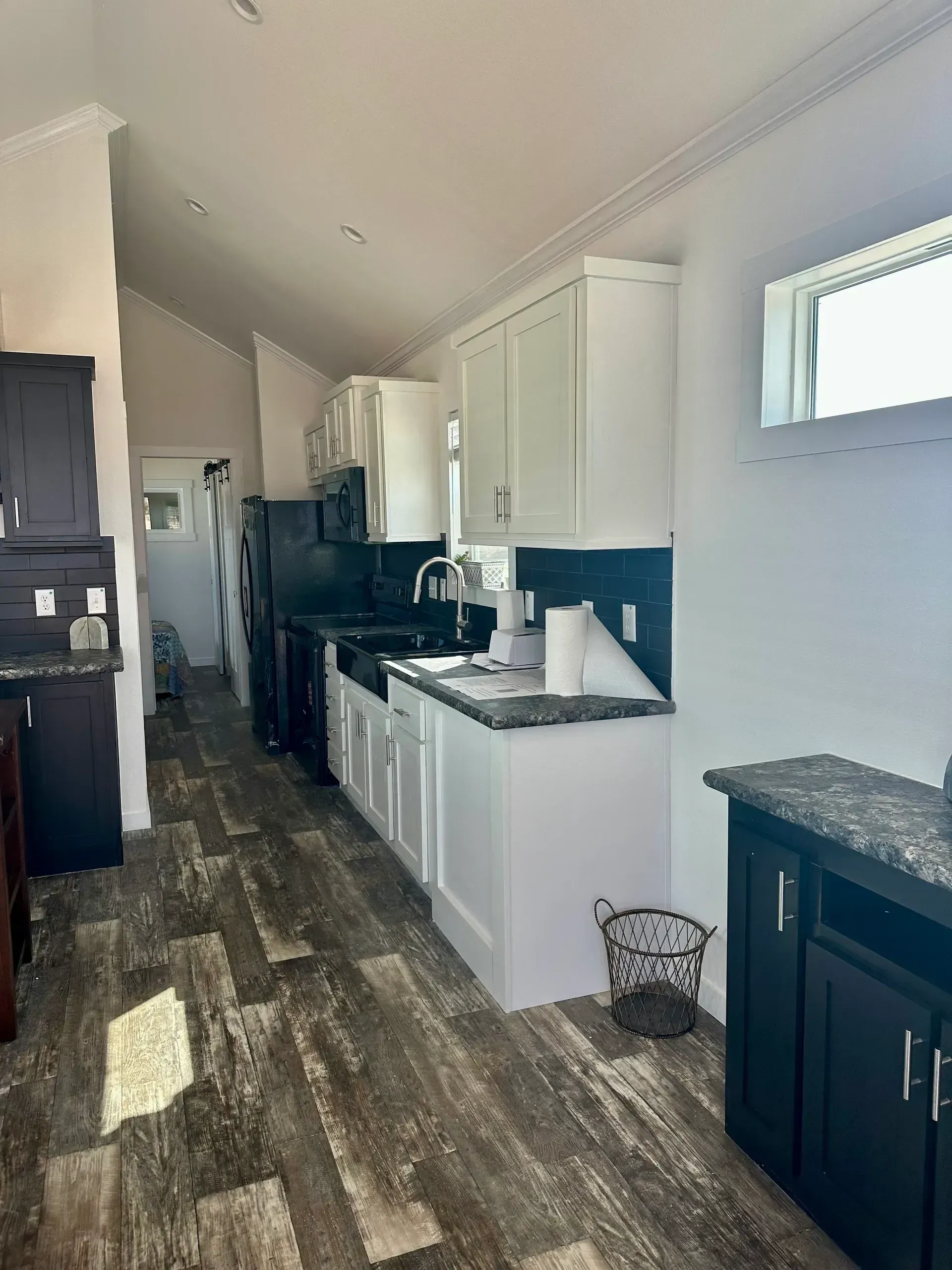 A narrow kitchen with white cabinets, dark countertops, and dark flooring, leading into an adjacent room.