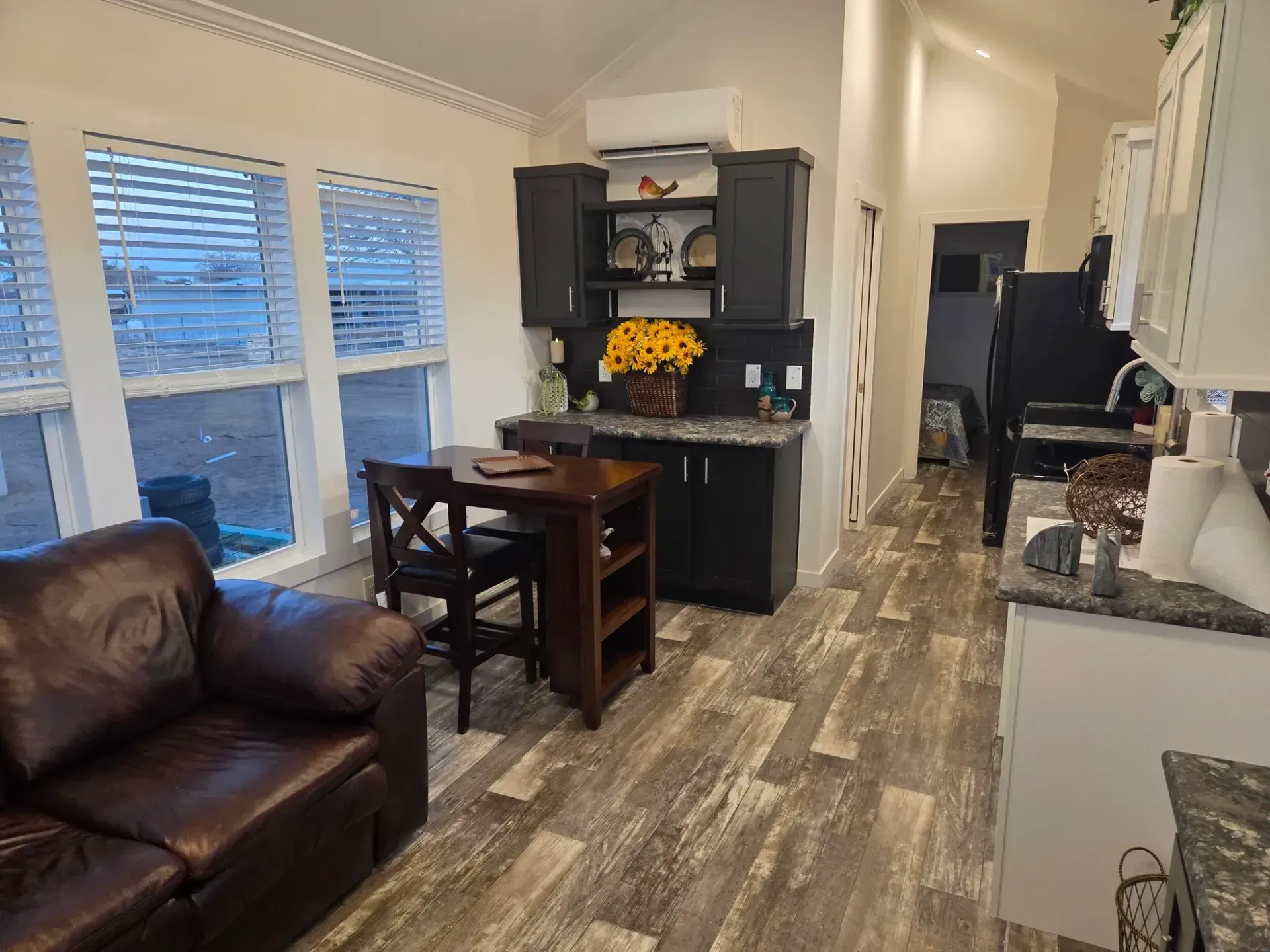 A cozy living area featuring a brown sofa, a small wooden dining set, dark kitchen cabinets, and light wood-plank flooring.