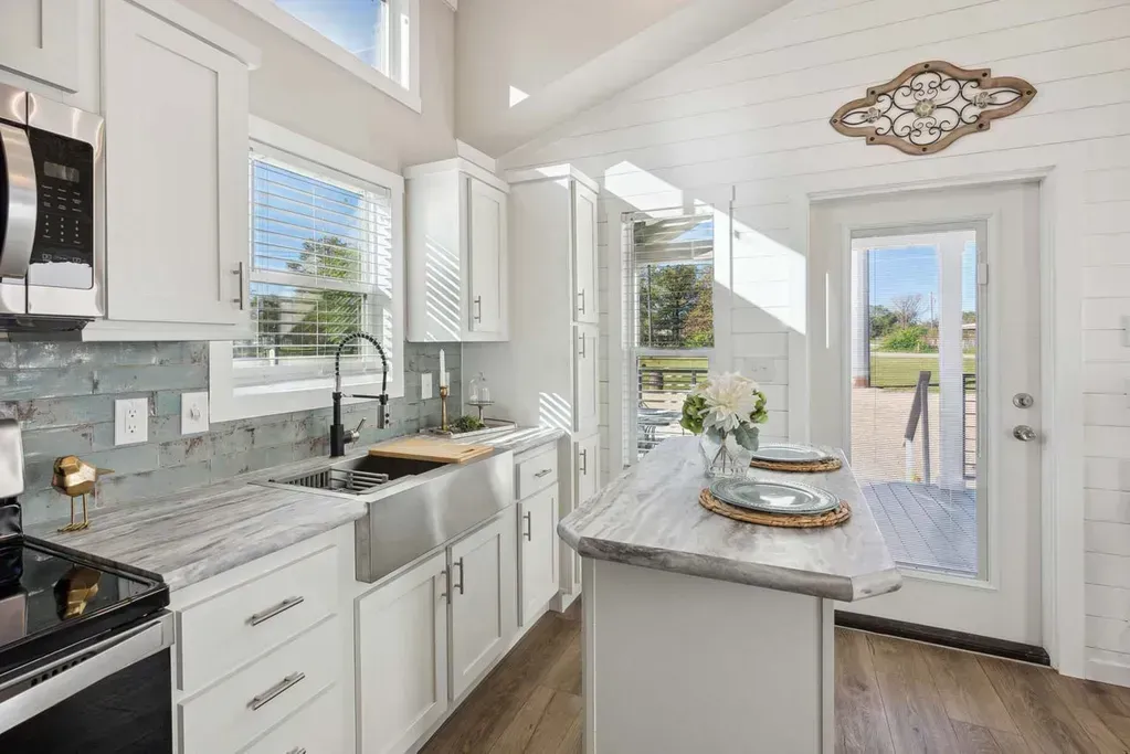 A bright, modern kitchen featuring white cabinets, stainless steel appliances, marble countertops, and a wood floor.