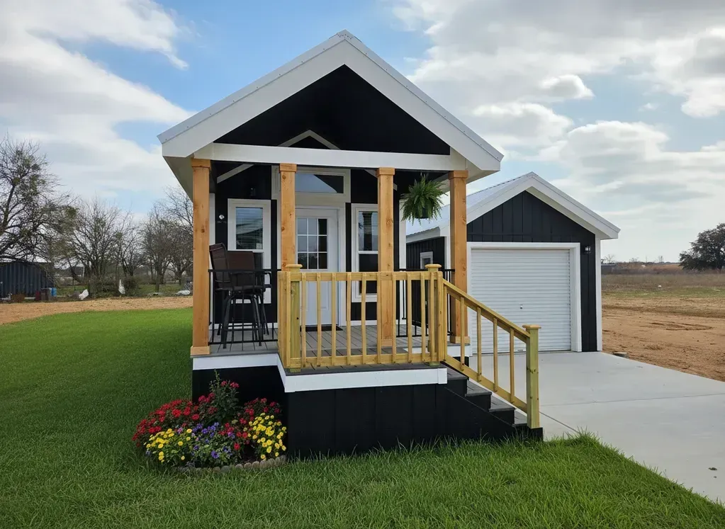 A black tiny house with white trim and a wooden front porch, featuring a small matching garage on a sunny, grassy lot.