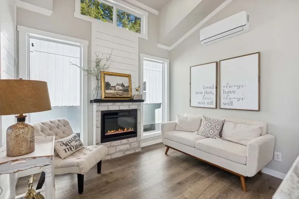 A living room with a fireplace, a white sofa, a tufted armchair, wood floors, and two framed prints on the wall.
