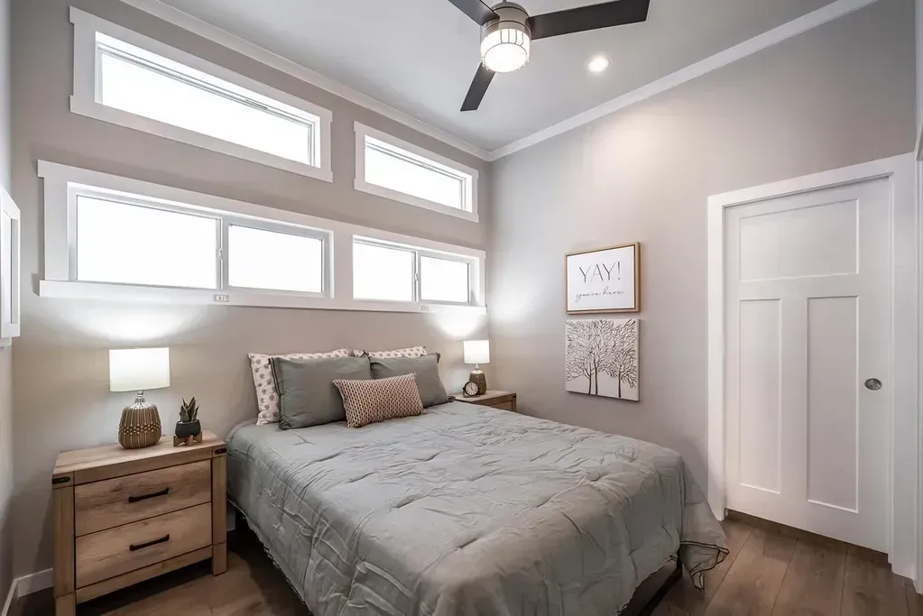 A bedroom with a queen bed, wooden nightstand, neutral bedding, ceiling fan, transom windows, and a white interior door.