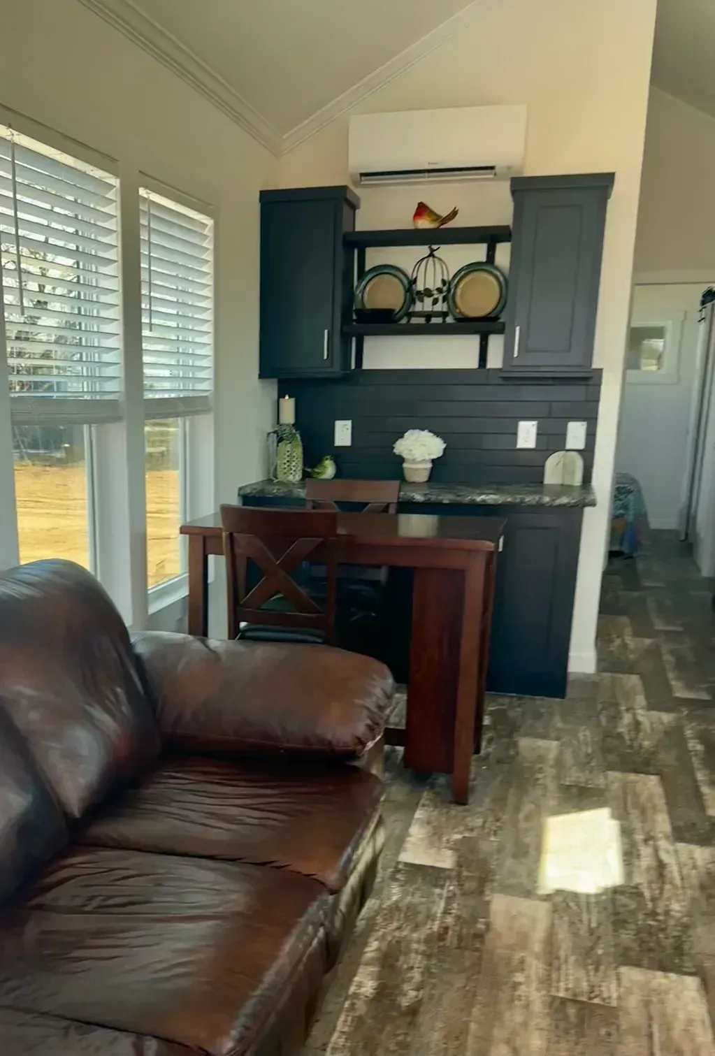 A dark brown leather sofa in the foreground faces a dining table, black cabinets, and an air conditioner in a living area.