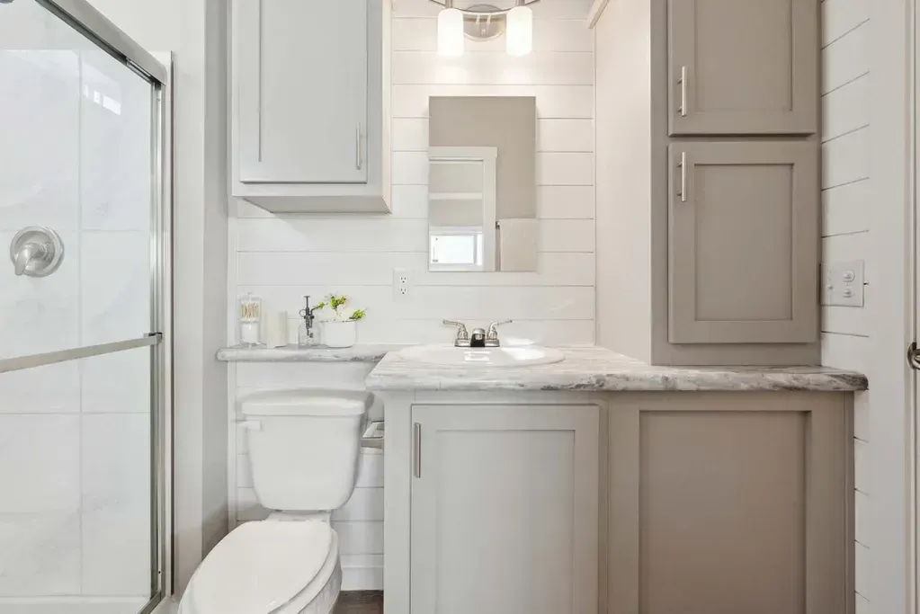 A modern bathroom featuring a white toilet, light gray vanity cabinets with a marble countertop, and a glass-door shower.