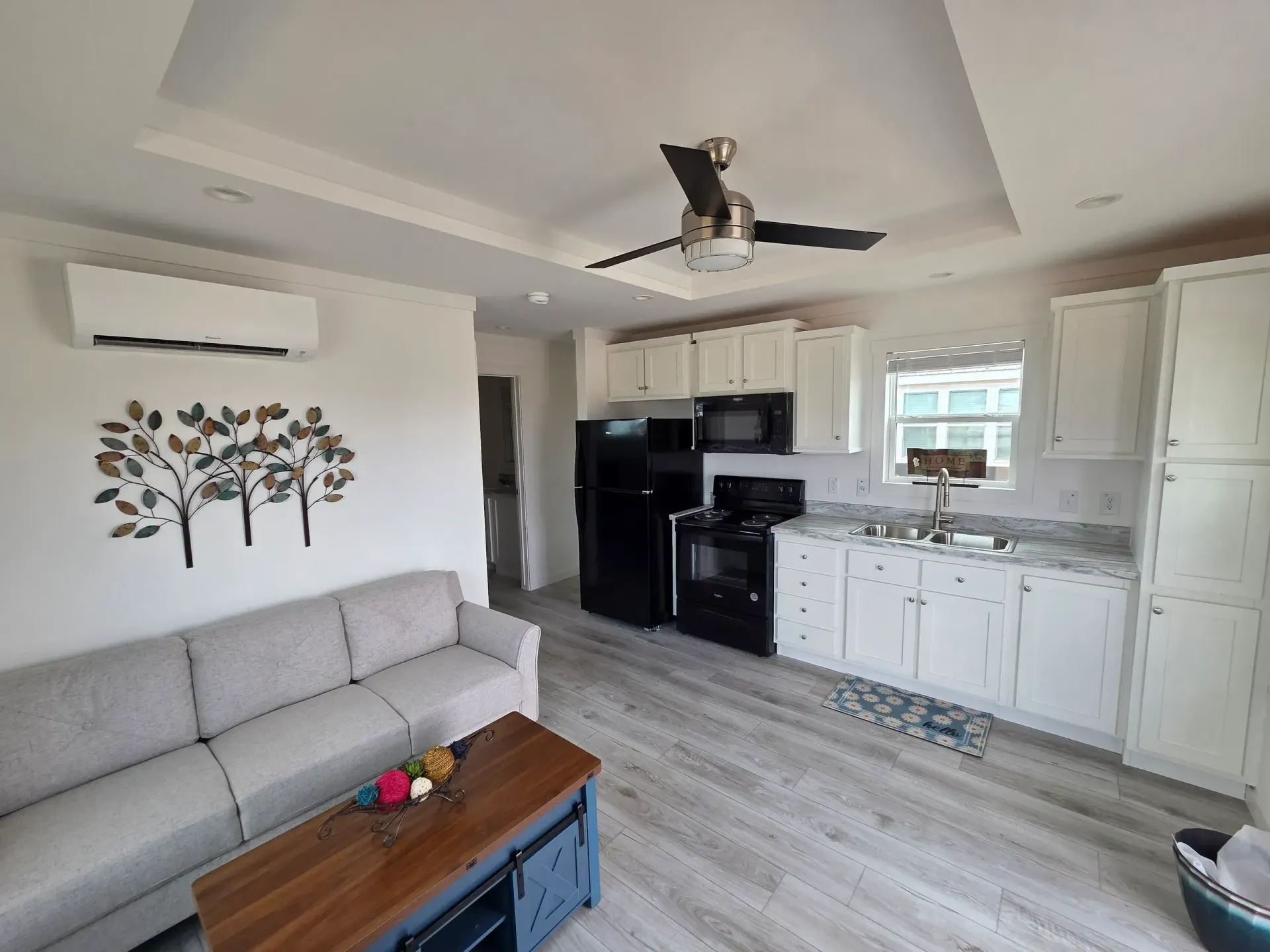 An open-concept living area with a light gray sofa, a wood coffee table, and a kitchen with white cabinets and appliances.