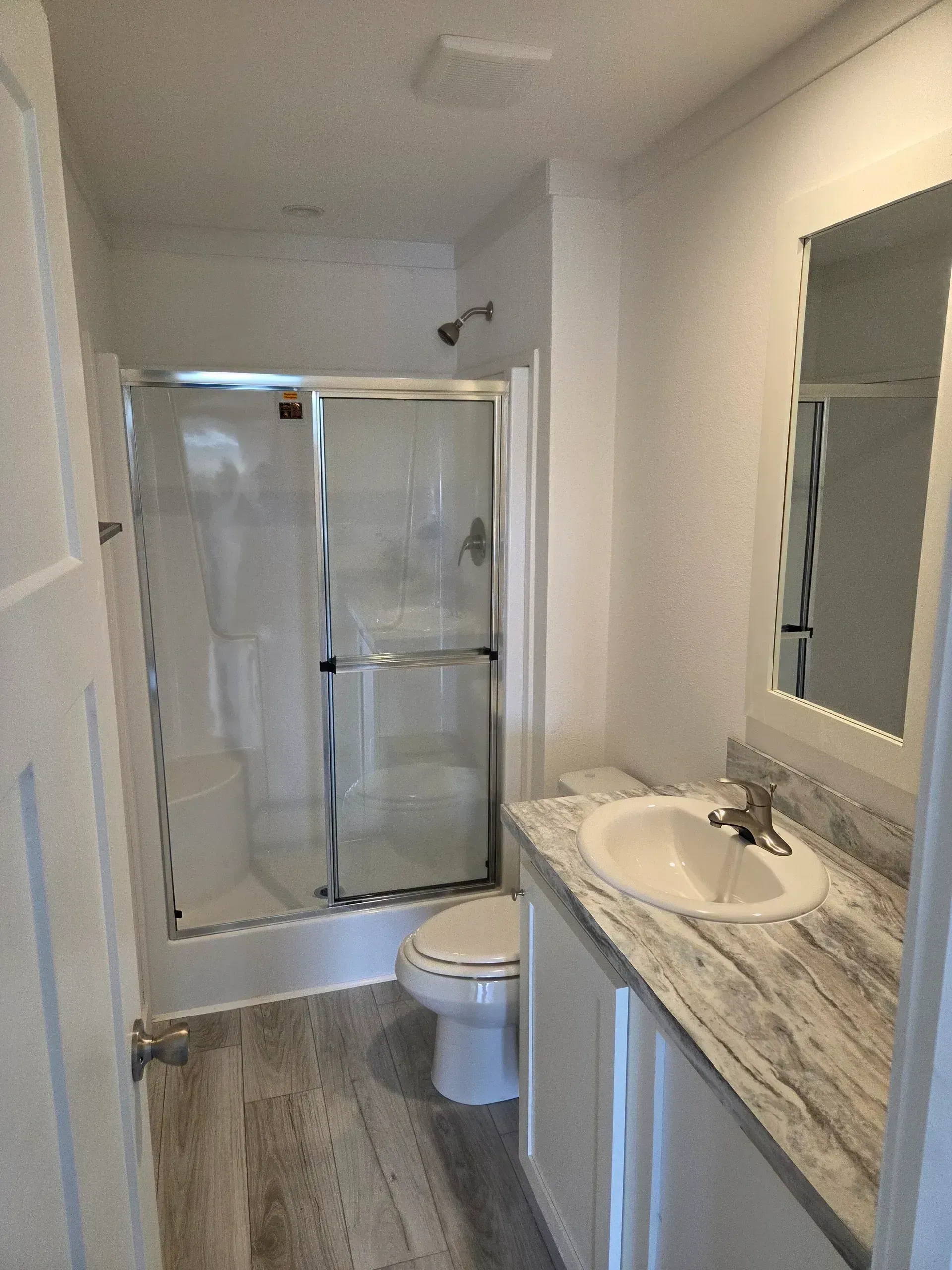 A bright bathroom with wood-look flooring, a white vanity with marble-patterned countertop, a toilet, and a glass shower.