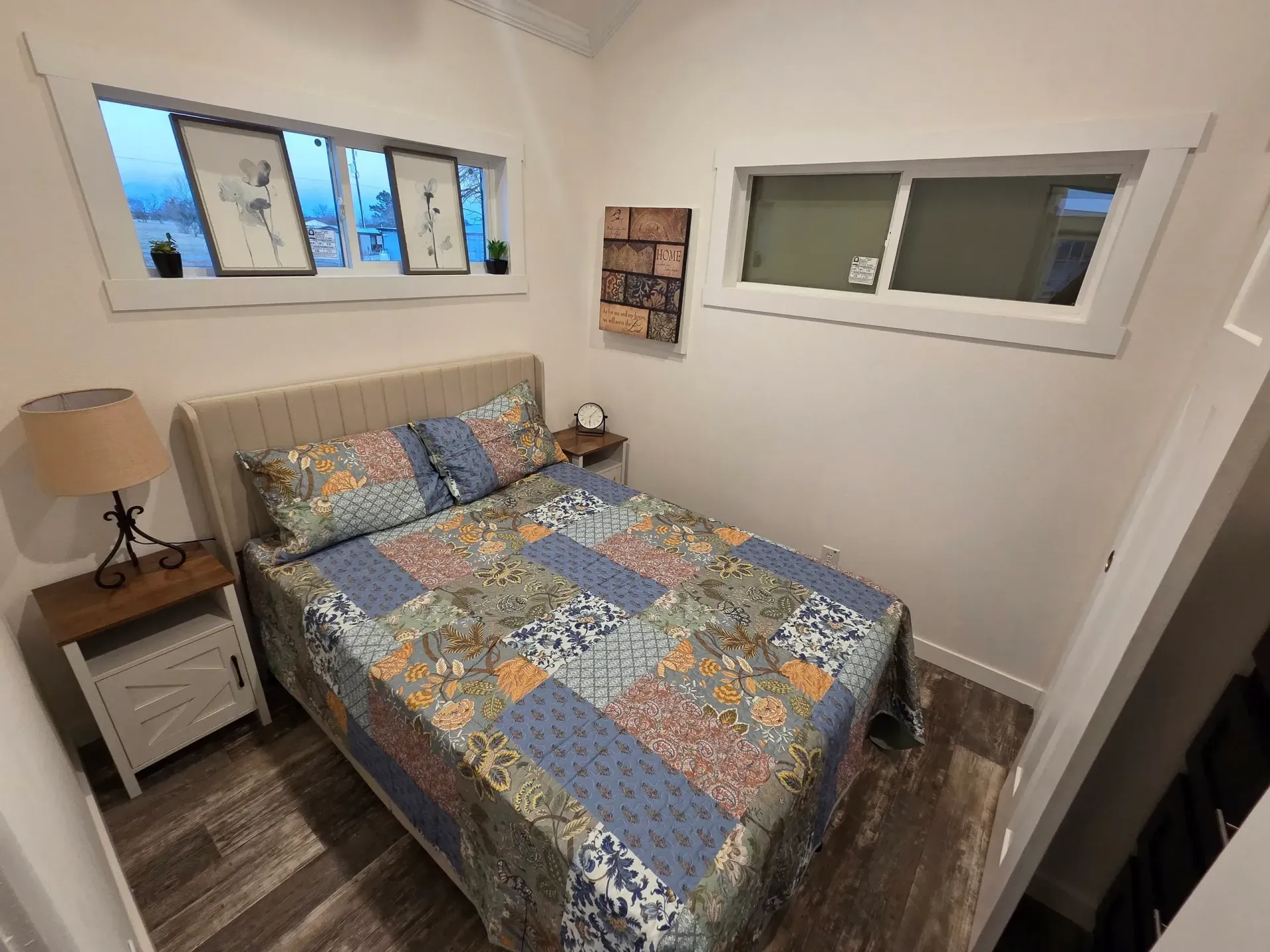 A small, bright bedroom featuring a queen bed with a blue floral patchwork quilt, a wood nightstand, and two wall windows.