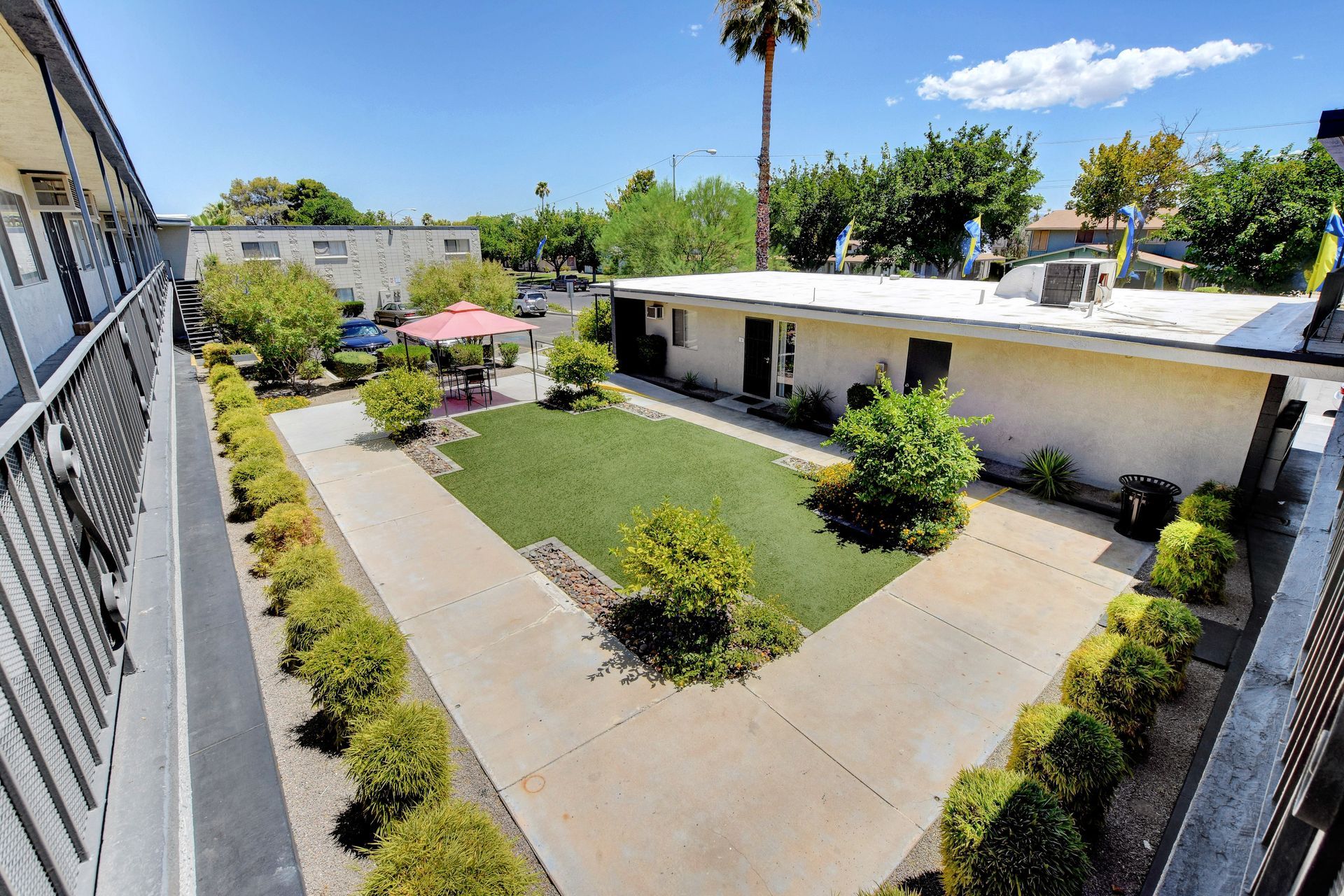 Apartment courtyard with white buildings, concrete paths, trimmed shrubs, and a grassy center under a sunny blue sky