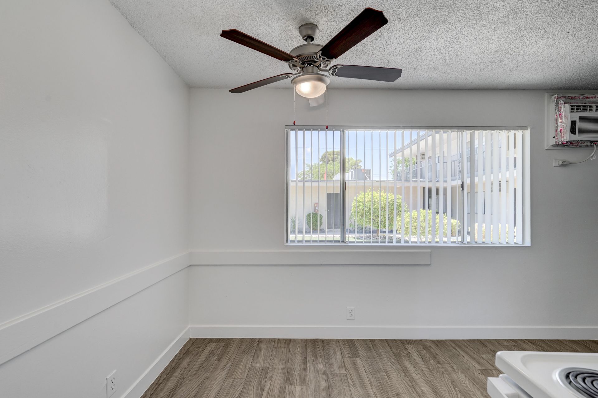 Empty white room with wood floor, ceiling fan, and window with vertical blinds.