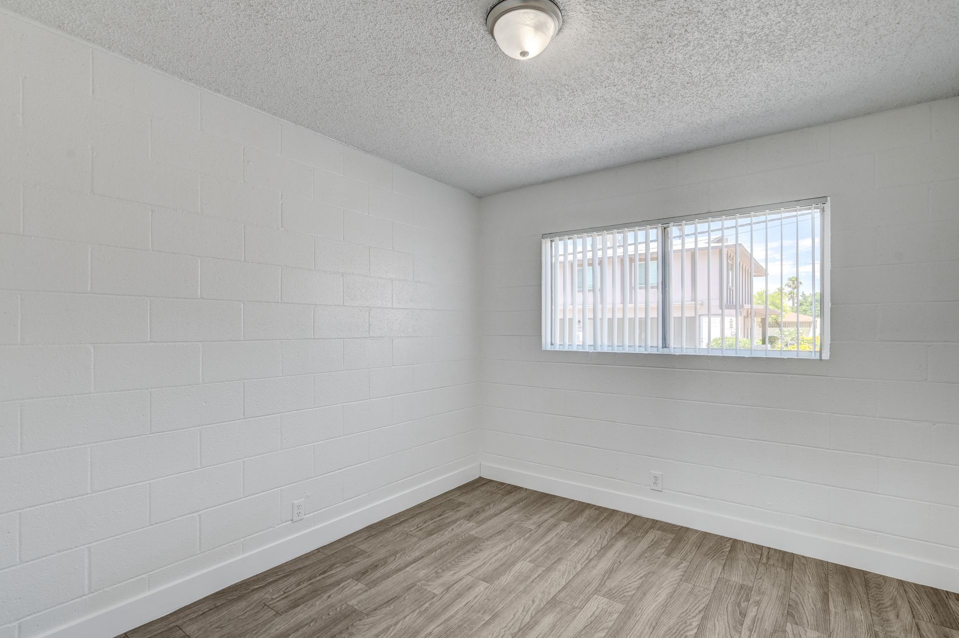 Empty white room with wood-look floor, cinderblock wall, and small window with blinds