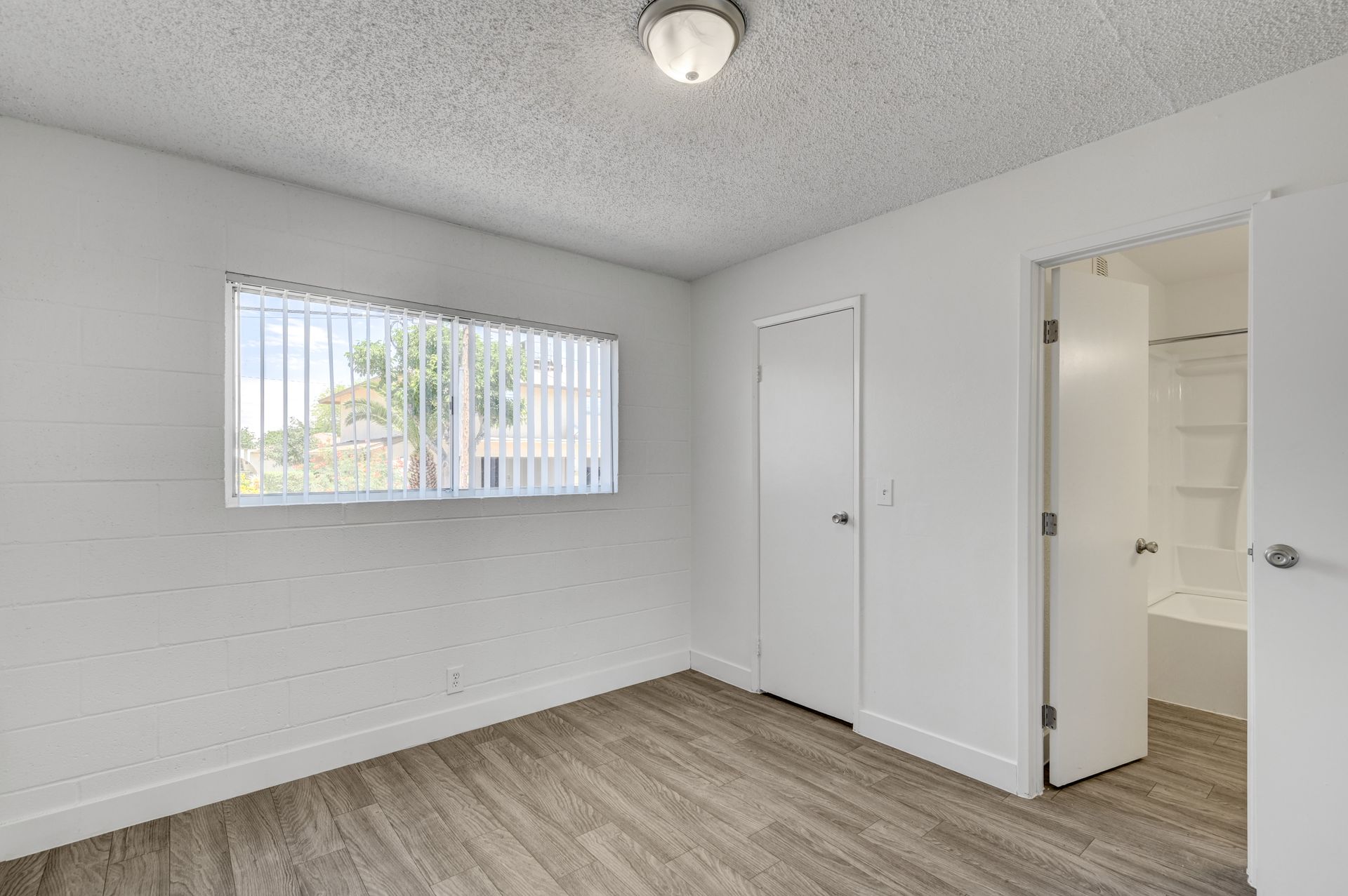Empty white bedroom with a window, closed closet door, and open bathroom door; wood-look flooring.
