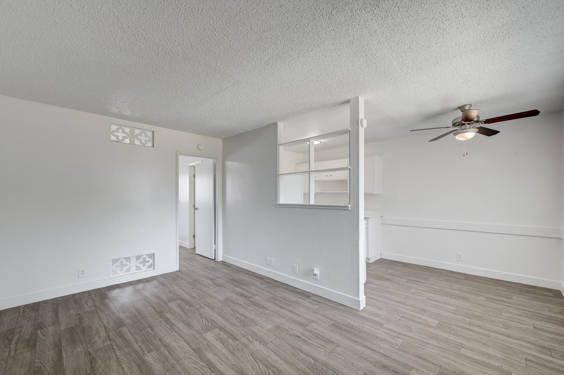 Empty light-gray living room with wood-look flooring, white trim, ceiling fan, and open doorway to a hall