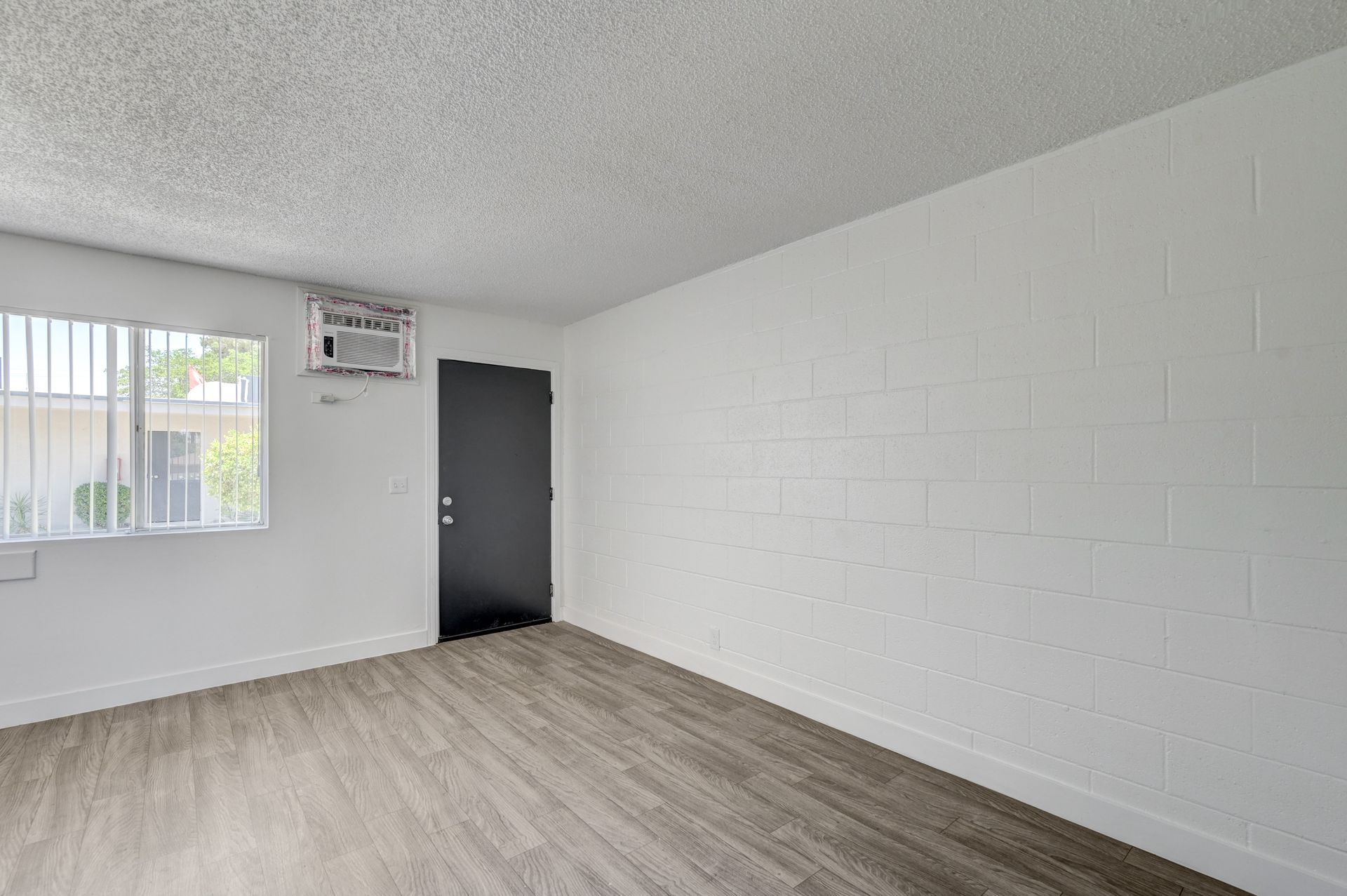 Empty room with white walls, wood-look floor, window, wall AC unit, and dark door.