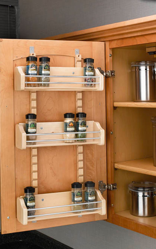 A wooden kitchen cabinet door with a mounted three-tier spice rack holding several glass spice jars.