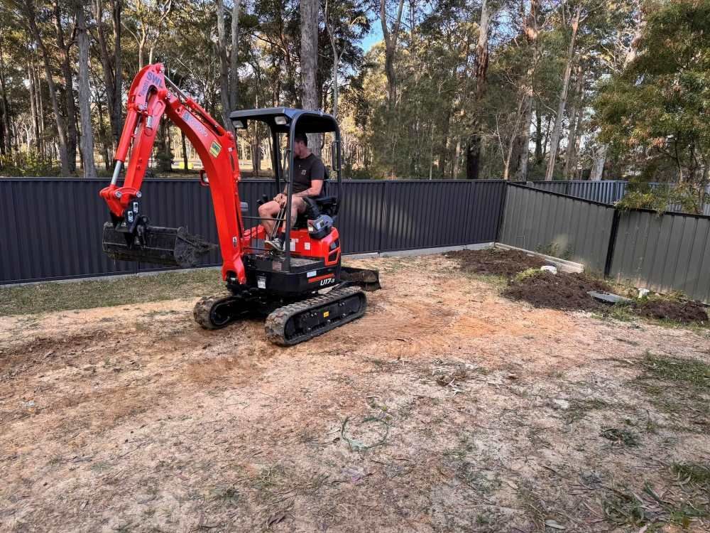 A Man is Driving a Small Red Excavator in a Backyard — Garrett's Excavation & Equipment Hire in Sanctuary Point, NSW