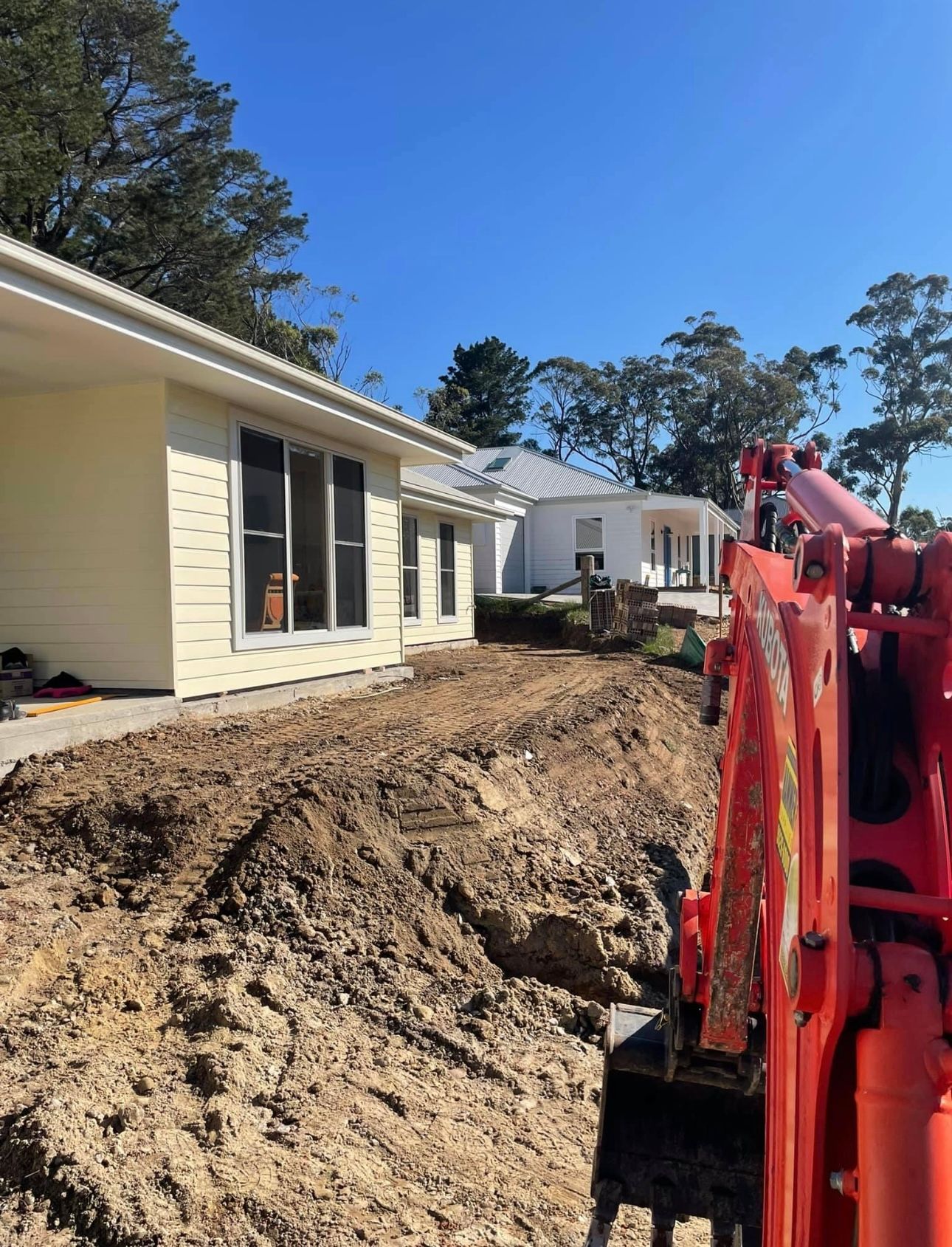 A Red Excavator is Digging a Hole in the Dirt in Front of a House — Garrett's Excavation & Equipment Hire in Sanctuary Point, NSW