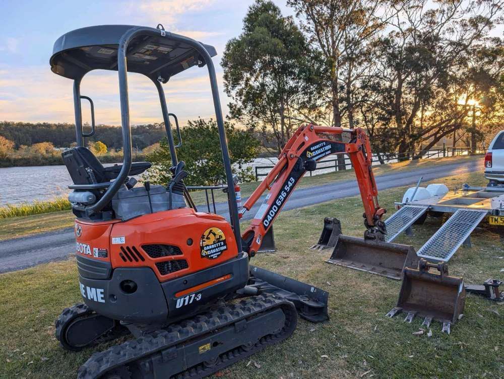An Excavator is Parked on the Grass Next to a Lake— Garrett's Excavation & Equipment Hire in Sanctuary Point, NSW