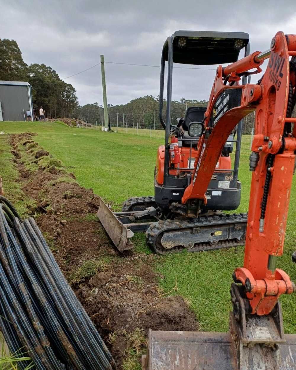An Orange Excavator is Digging a Hole in a Grassy Field — Garrett's Excavation & Equipment Hire in Saint Georges Basin, NSW