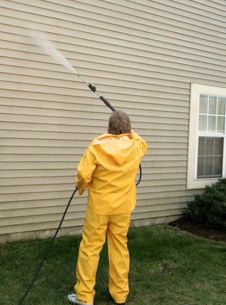 Person in yellow rain gear pressure-washing house siding in a yard