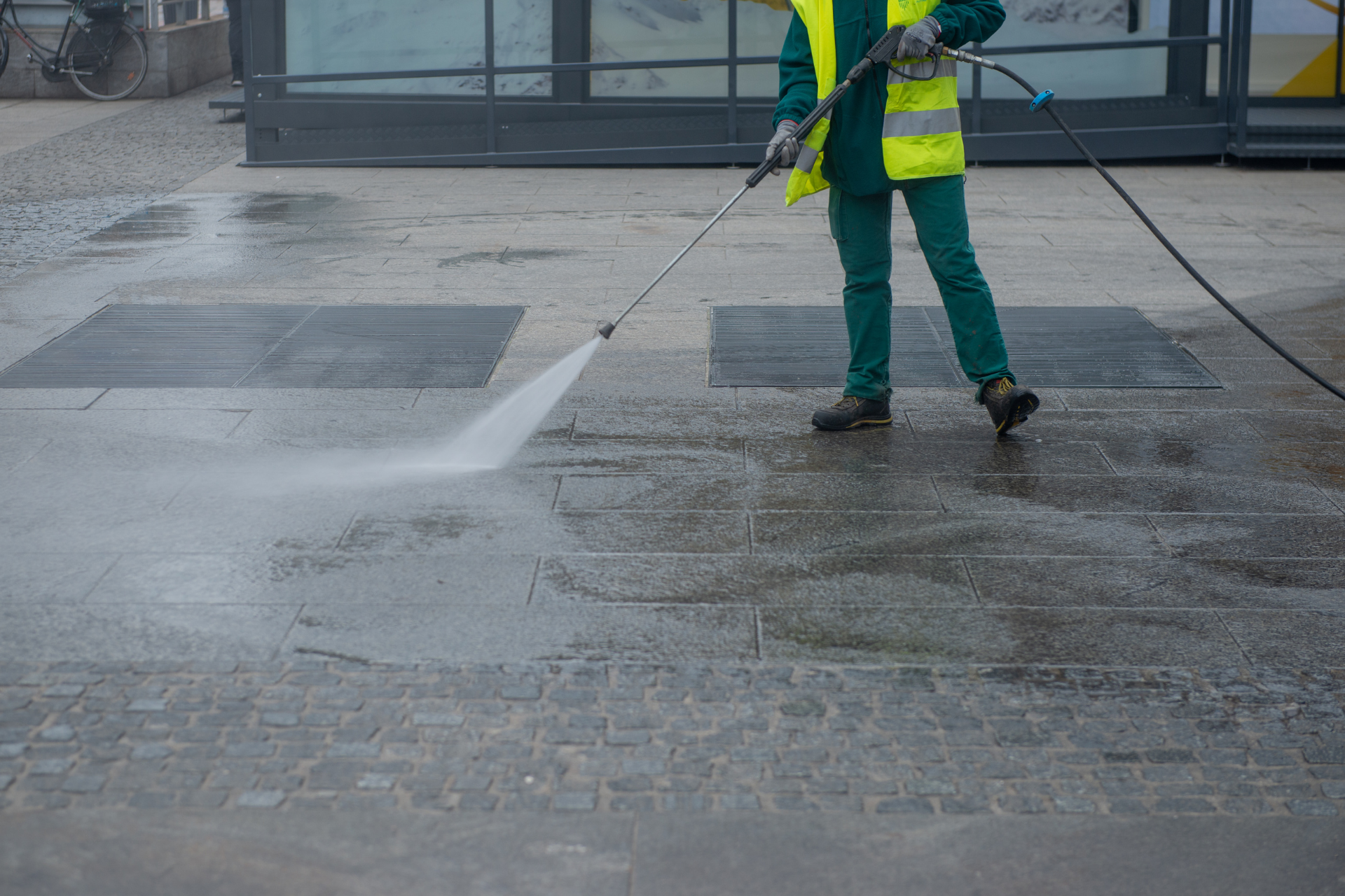 Worker in yellow vest pressure-washing a wet sidewalk outside a building