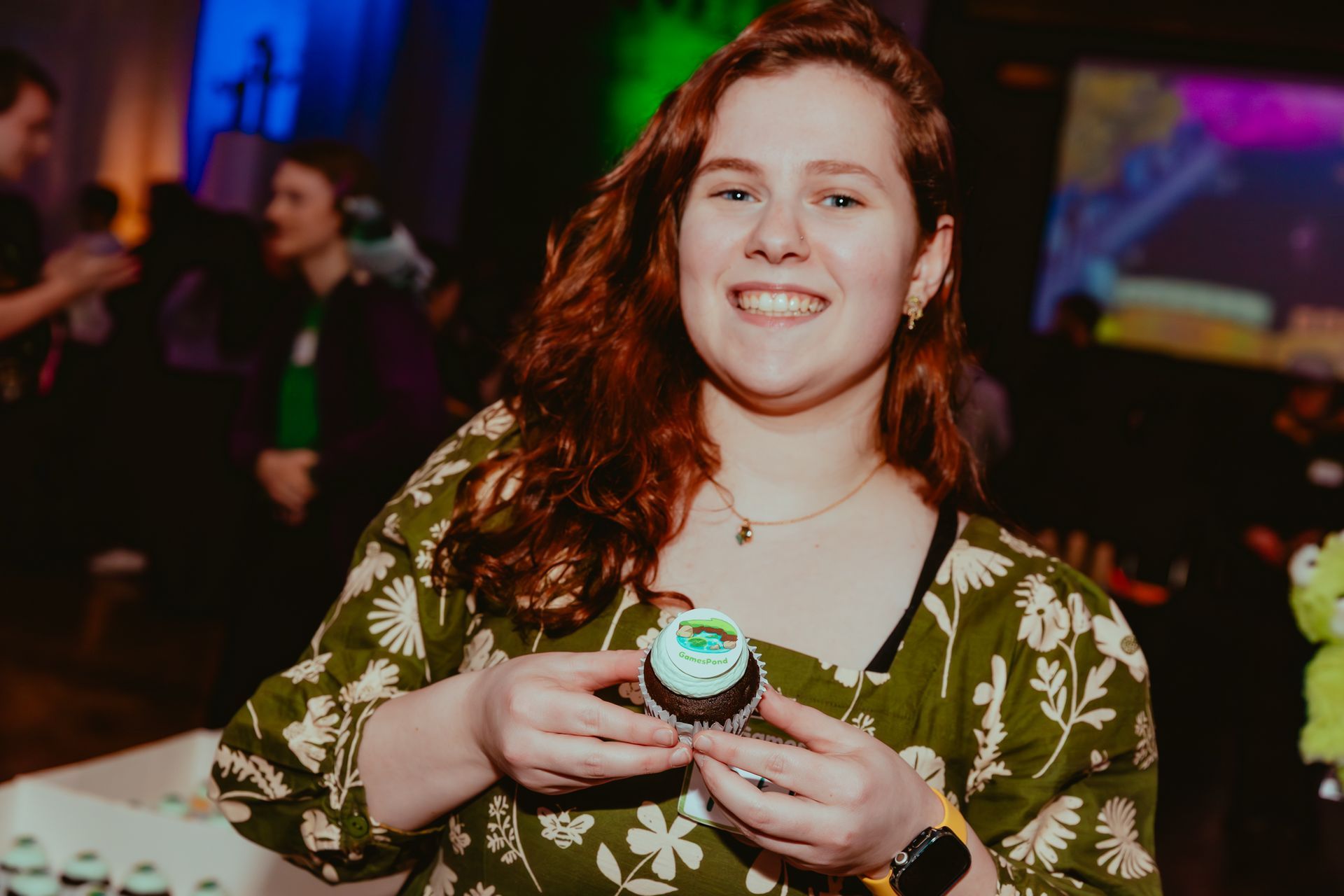 A woman in a yellow sweater is smiling in front of a green hedge.