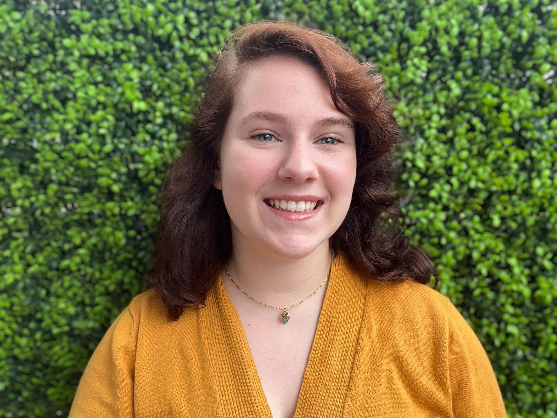 A woman in a yellow sweater is smiling in front of a green hedge.