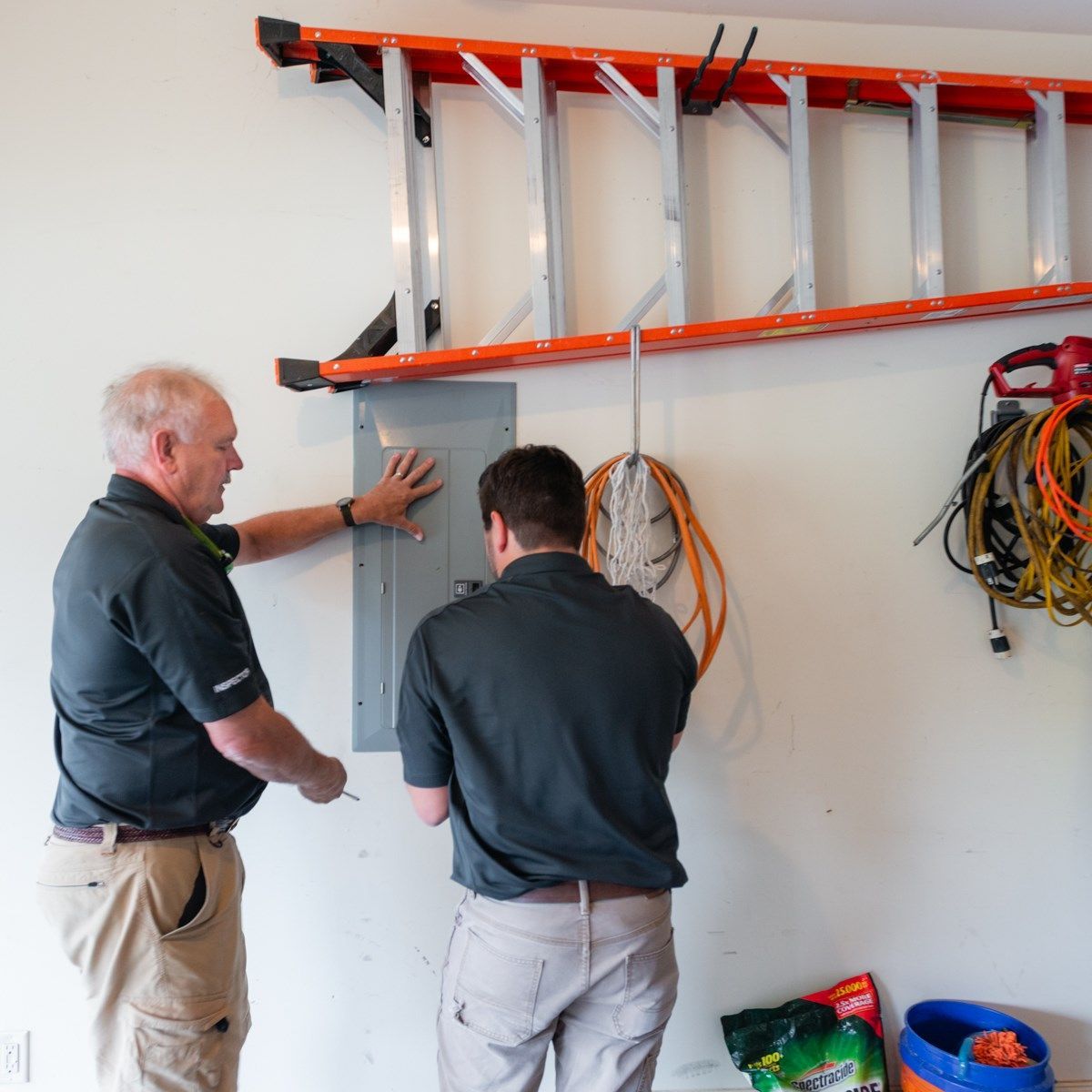 Two men inspecting electrical panel in a garage, ladder and wires visible.