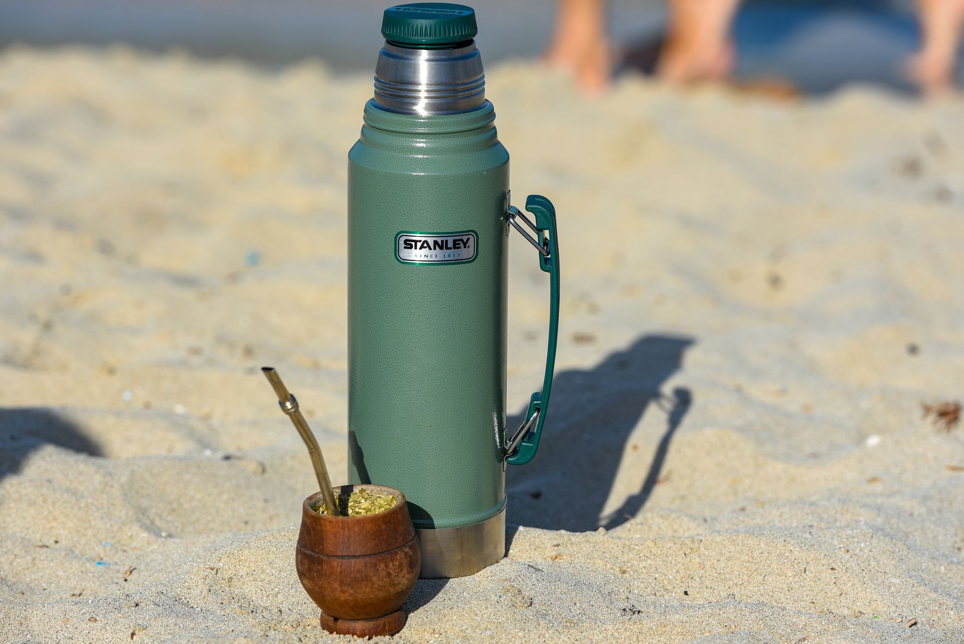 Termo Green Stanley y taza de mate en una playa de arena.