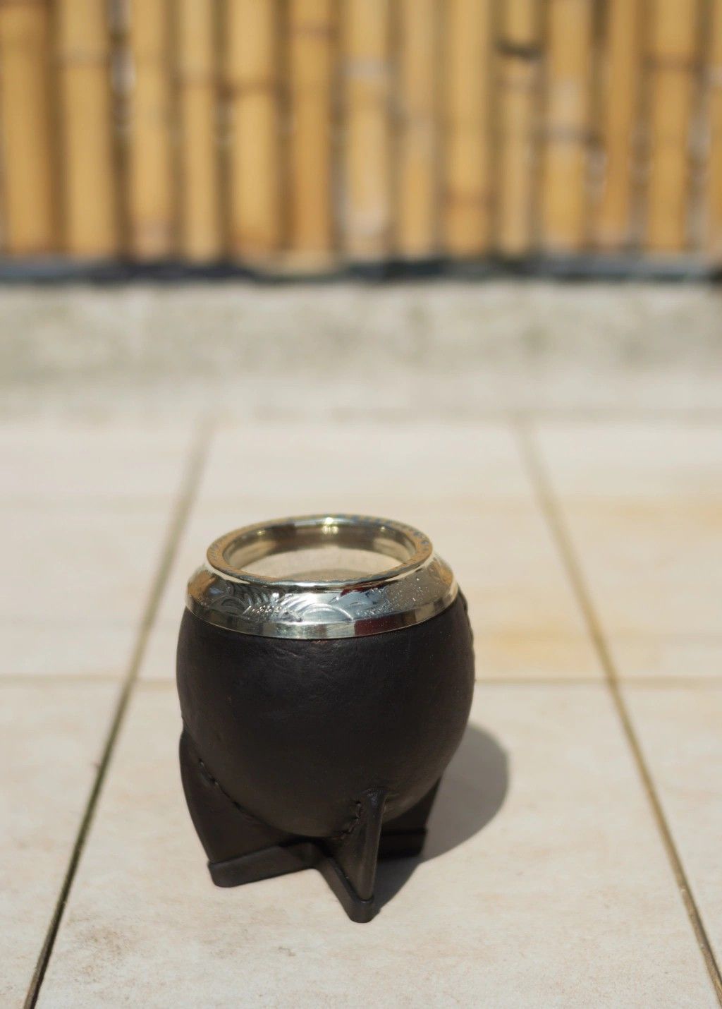 Una calabaza negra con borde plateado, apoyada sobre una superficie de baldosas con un fondo de bambú.
